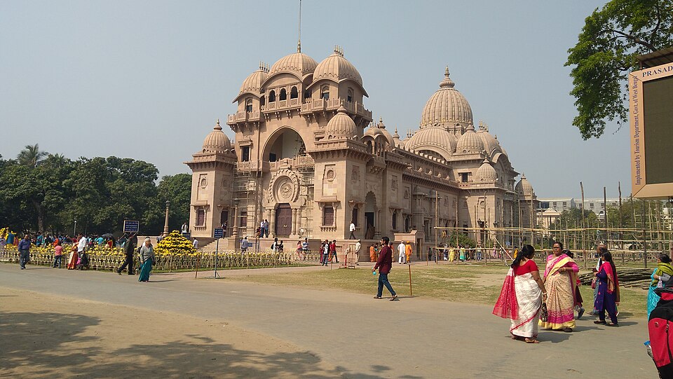 The Sri Ramakrishna Temple at Belur Math in full daylight: a long sandstone-coloured building crowned by a cluster of ribbed domes and a tall central arch that mixes temple, mosque and church forms, with beds of marigolds and visitors walking on the open ground in front of it.