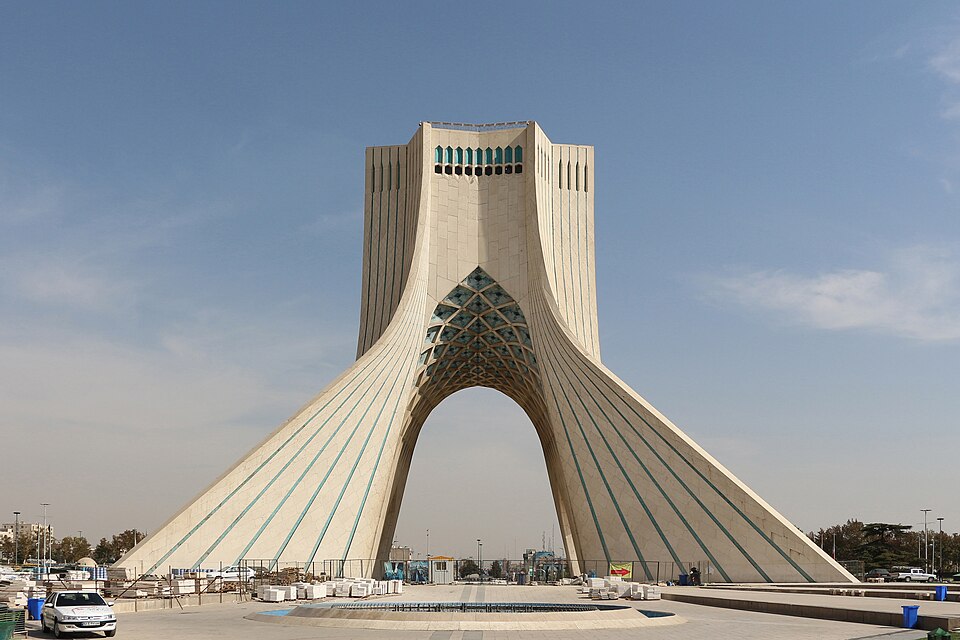 The Azadi Tower in Tehran seen square-on in daylight: a white marble monument whose two curving legs sweep up off the plaza and meet in a tall pointed arch, the underside of the arch filled with a turquoise-tiled net vault and a band of turquoise grillework set high in the block above, with a paved square, a low pool, stacked building materials and a parked white car along the bottom of the frame under a pale sky.