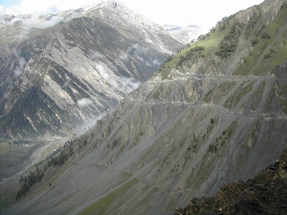 The Zoji La road seen from above: an unsurfaced single-lane shelf cut straight into an enormous grey scree slope, doubling back on itself in three visible tiers as it descends, with a green valley floor and scattered settlement far below at the left, cloud snagged on the bare rock face opposite, and snow lying on the ridges at the head of the valley.