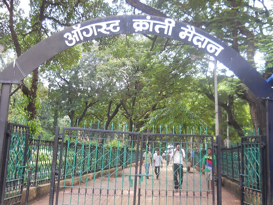 The entrance to August Kranti Maidan in Mumbai: a black steel arch carrying the name in large white Devanagari letters spans a pair of open green wrought-iron gates, with a paved path leading into a green, heavily treed park and a few people walking out.