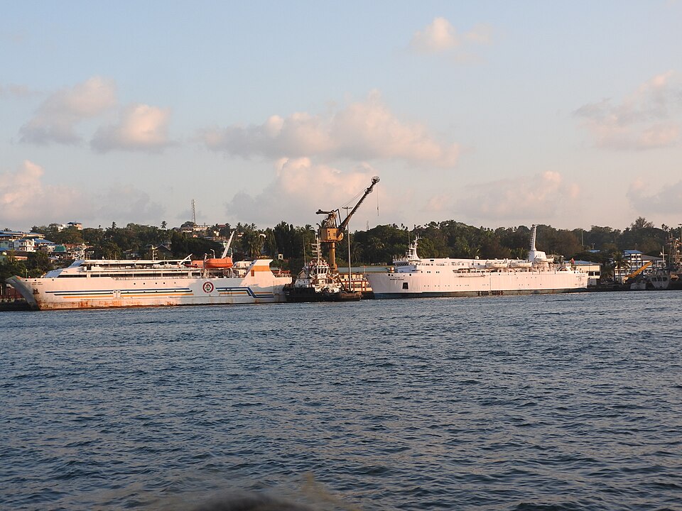 Port Blair harbour in evening light seen across choppy blue water: two large white passenger ships lie alongside Haddo wharf with a yellow dockside crane between them, and behind runs a low green shore of palms and scattered houses under scattered cumulus.
