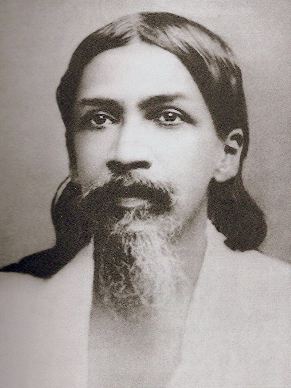 A sepia studio portrait of Sri Aurobindo facing the camera: long dark hair falling past his shoulders, a heavy dark moustache and a long beard going white at the chin, calm steady eyes, wearing a plain light wrap against a blank pale background.