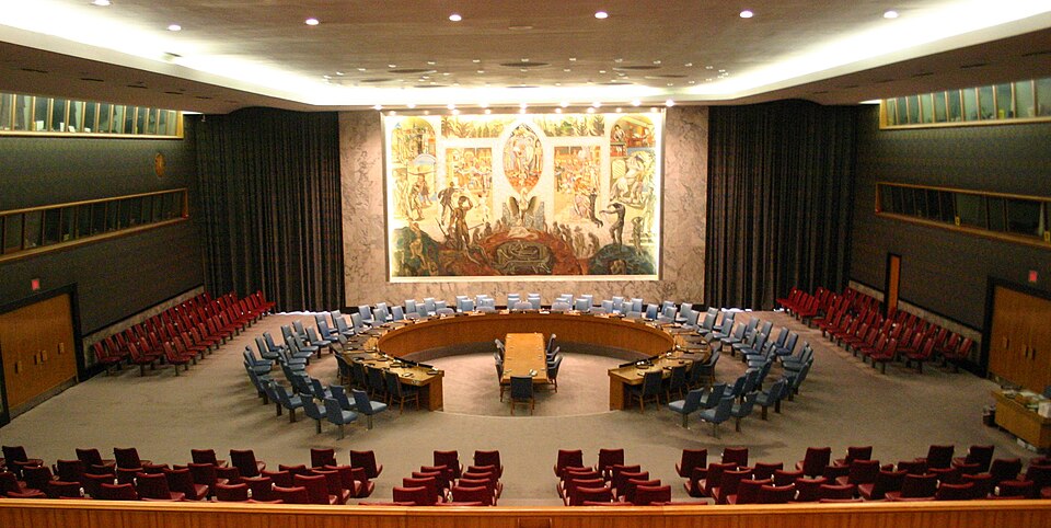 The empty United Nations Security Council chamber: a large horseshoe-shaped wooden table ringed with blue chairs on a raised floor, banks of red seats for delegations and the public around it, and the Per Krohg mural of a phoenix rising from ashes filling the wall behind.