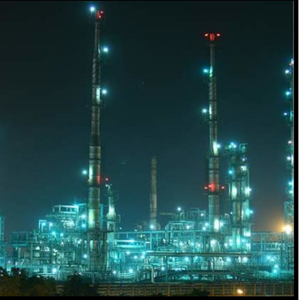 The Barauni oil refinery at night: two tall slender distillation columns rise against a black sky, and the plant below them — a dense tangle of pipework, vessels and gantries — is picked out in thousands of blue-white lamps, with red aircraft-warning lights on the towers.