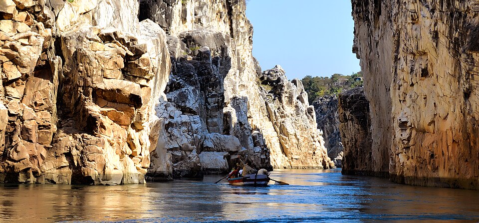 The Narmada squeezed into the Bhedaghat gorge: sheer, sunlit cliffs of cream and honey-coloured marble rise straight out of the water on both sides, and a single small wooden rowing boat with two people in it sits mid-channel, dwarfed by the walls.