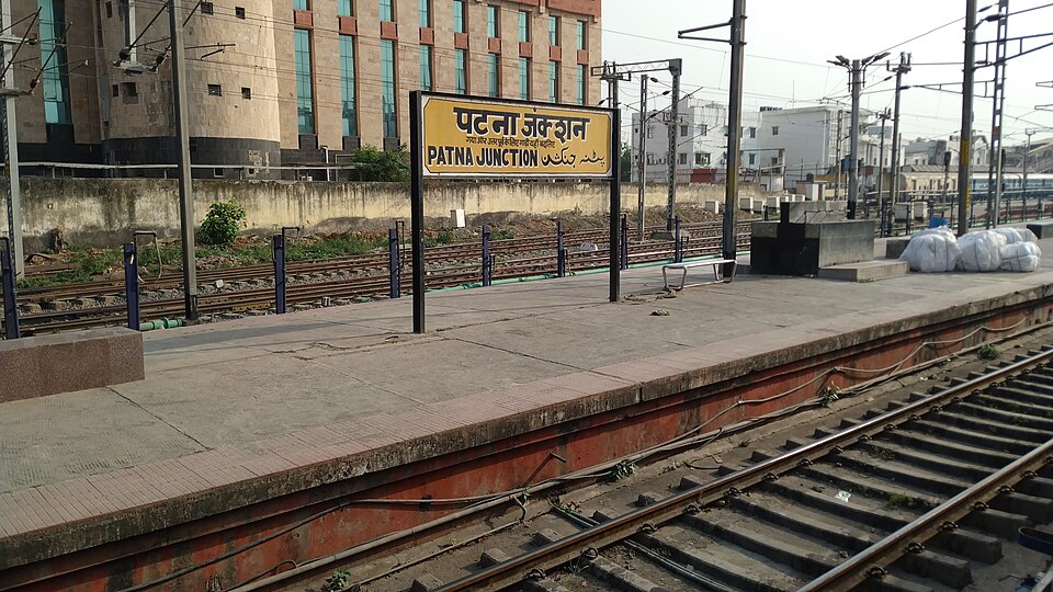 An empty platform at Patna Junction with a yellow station board on black poles reading the name in Hindi, English and Urdu; bare tracks run along both sides, overhead electrification masts stretch into the distance, and a few white bundles of luggage sit at the far end.