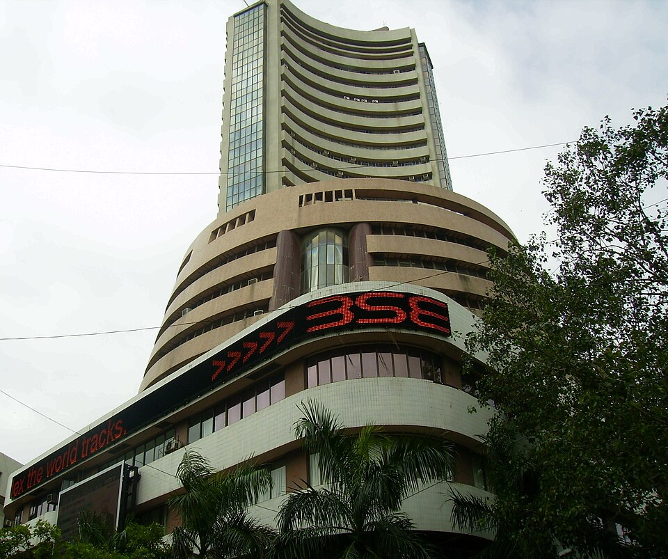 Looking up at Phiroze Jeejeebhoy Towers, the Bombay Stock Exchange building: a tall tower of stacked curved concrete balconies above a rounded podium, with a long red LED ticker board wrapping around the facade and a strip of advertising reading 'the world tracks' at street level, palms in the foreground.