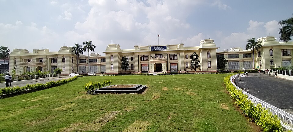 A straight-on daylight view of the Bihar Legislative Assembly in Patna: a low, symmetrical cream building with a pillared central porch, arched windows and a blue signboard above the entrance reading the name in Hindi and English, fronted by a wide mown lawn and clipped hedges.