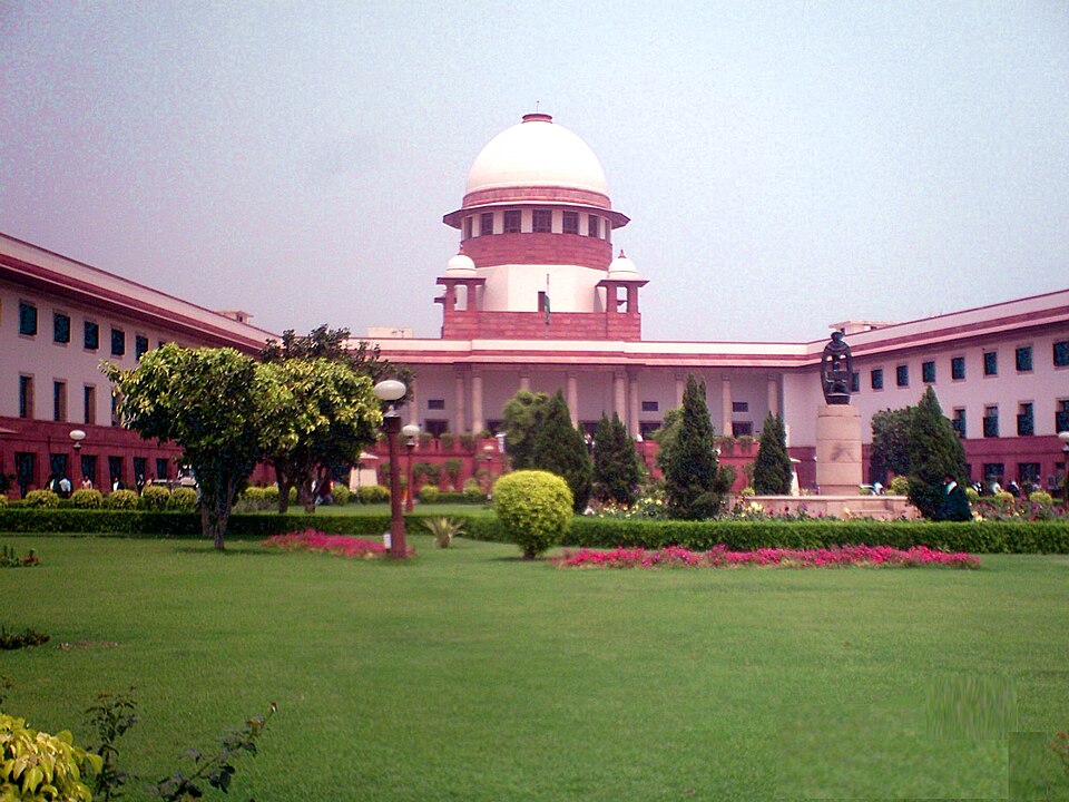 The Supreme Court of India seen across its front lawn: a white ribbed dome on a red-and-cream drum flanked by two small chhatris, rising above long low wings of red sandstone and white plaster, with a dark bronze statue on a plinth to the right and beds of pink flowers on the grass.