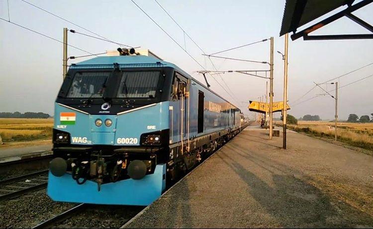 A twin-section blue-and-white WAG-12 electric locomotive numbered 60020, tricolour roundel on its nose and pantograph raised to the overhead wires, running through a small rural platform at dawn with fields on either side