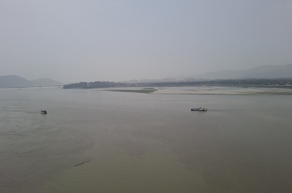 The Brahmaputra spread out wide and brown at Guwahati on a hazy day, two small ferries crossing mid-stream, a long pale sandbar dividing the channel and the low forested hills of the far bank with a road bridge on the left