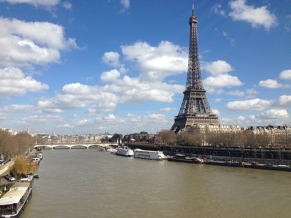 The Seine curving through Paris under a blue sky of scattered cloud, with the Eiffel Tower rising on the right bank above a row of Haussmann apartment blocks, white cruise boats and barges moored along the stone quays and an arched bridge in the distance