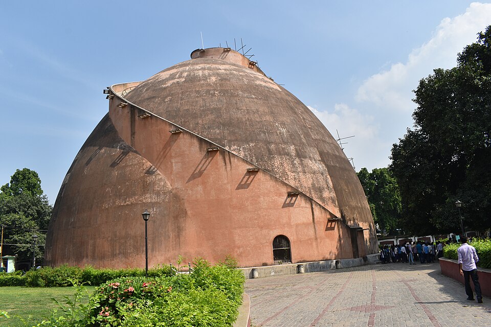 The Golghar at Patna — a huge beehive-shaped dome of pinkish plastered brick with a small arched doorway at its base and an external stairway spiralling up one flank to an opening at the crown, standing in a lawned park with visitors on the paved path beside it