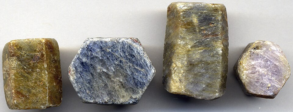 Four rough corundum crystals laid in a row on a grey surface — stubby barrel-shaped prisms with flat hexagonal ends, ranging from dull olive-brown through steel blue to pale lilac, uncut and unpolished
