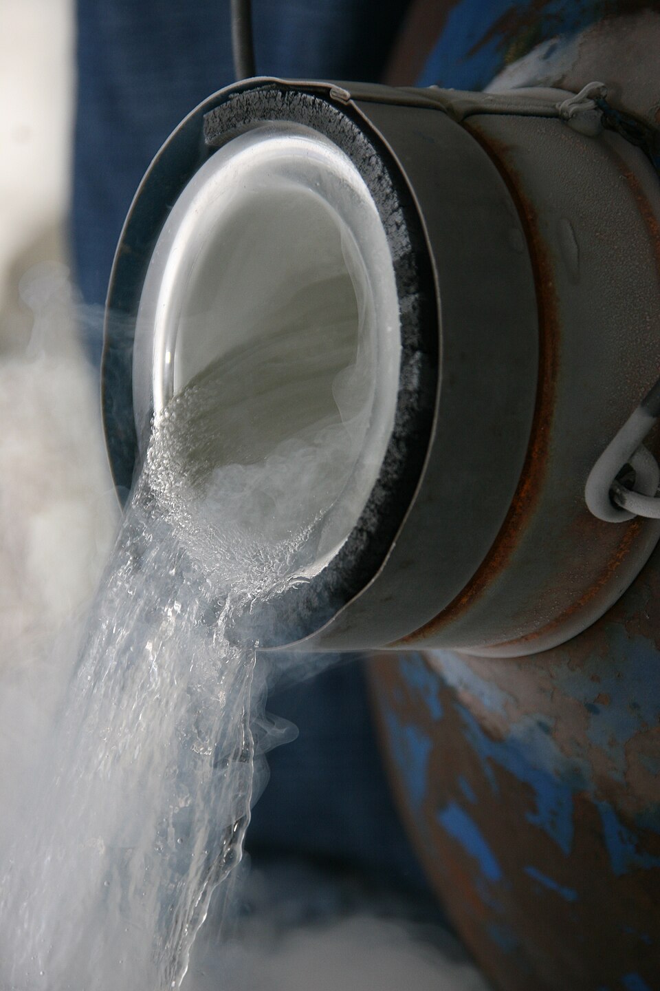 Close-up of liquid nitrogen being tipped out of a metal cryogenic flask whose rim is furred with white frost — a clear, boiling stream falls from the lip and thick white condensation vapour rolls away from it across the frame