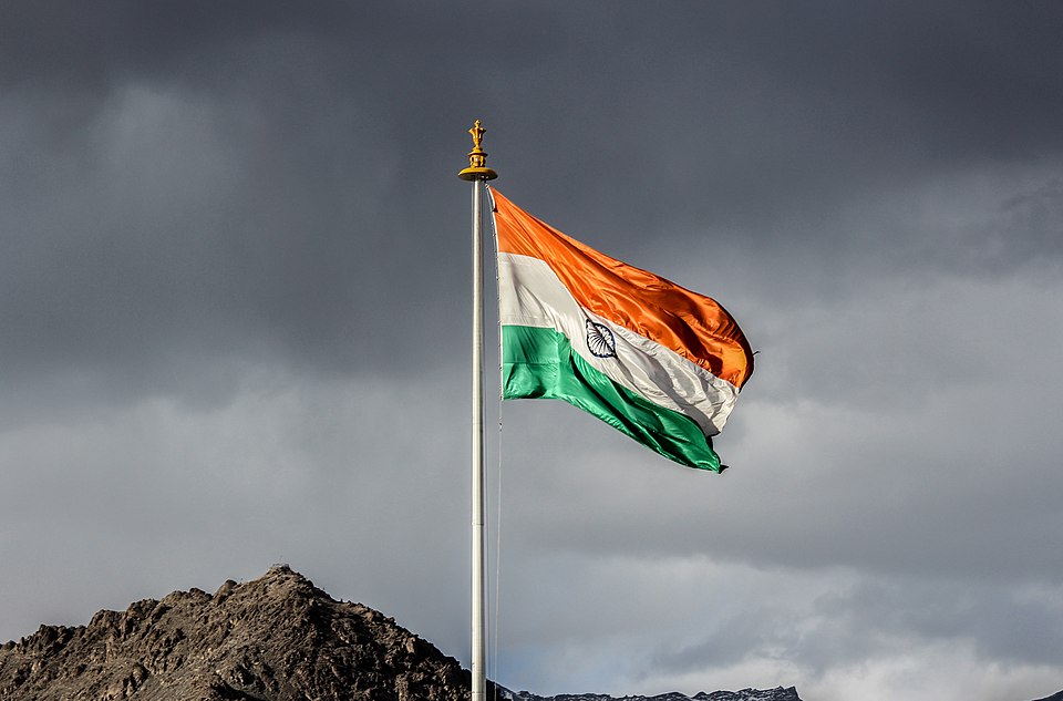 The Indian tricolour streaming from a tall metal flagpole topped with a gold Ashoka-lion finial, the navy-blue spoked chakra clearly visible in the white band, against a bank of dark storm cloud with a bare rocky ridge below