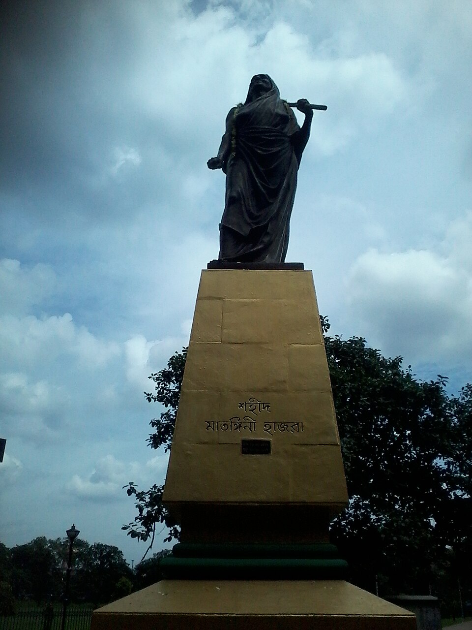 A dark bronze statue of a sari-clad woman striding forward with one arm raised over her shoulder holding a broken staff, mounted high on a tall tapering sandstone plinth inscribed in Bengali 'Shahid Matangini Hazra', seen against a cloudy sky with trees behind