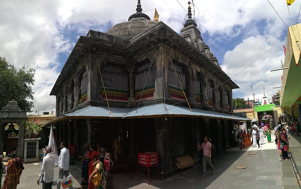 गया के विष्णुपद मंदिर का गहरे धूसर पत्थर वाला मंडप — जालीदार झरोखों वाला स्तंभयुक्त कक्ष, ऊपर सीढ़ीदार पिरामिडी छत, पीछे उठता सुनहरे कलश वाला धारीदार शिखर, ऊपर तनी रस्सियाँ और केसरिया पताकाएँ, और पक्के आँगन से गुज़रते तीर्थयात्री