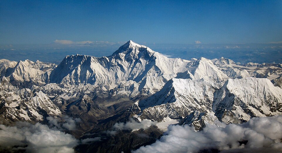 An aerial view along the Himalayan crest: the dark rock pyramid of Everest standing above a sea of snow-plastered ridges, Lhotse's white summit rising immediately to its right beyond the saddle that links them, hazy blue ranges behind and cloud banked in the valleys below