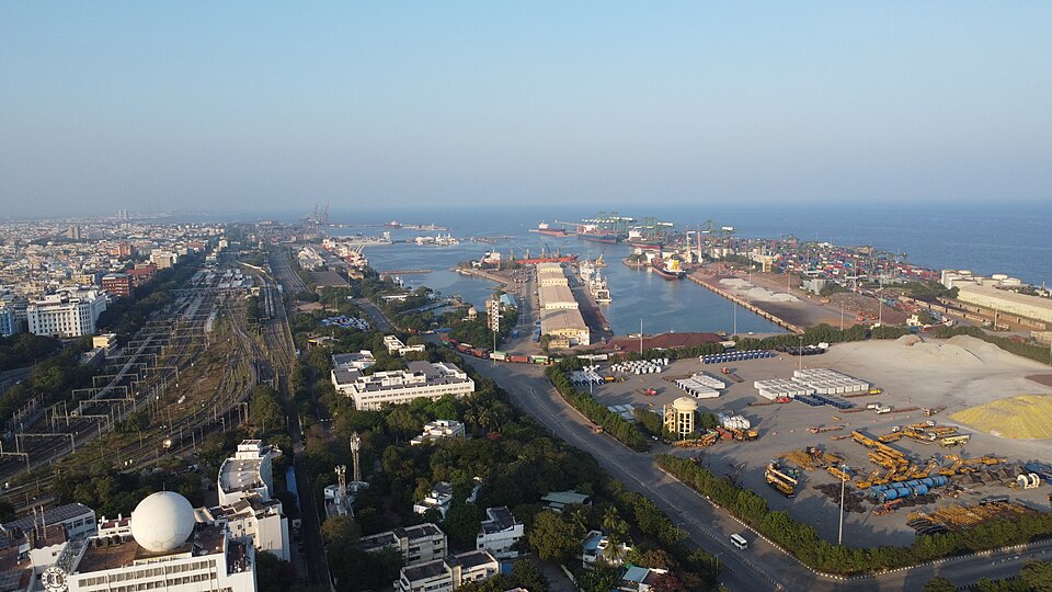 An aerial view over Chennai Port — quays, gantry cranes and stacked containers on made ground beside the city's rail yard, with long straight breakwater arms running out into the Bay of Bengal to enclose a rectangle of still water where ships lie berthed, and open sea beyond