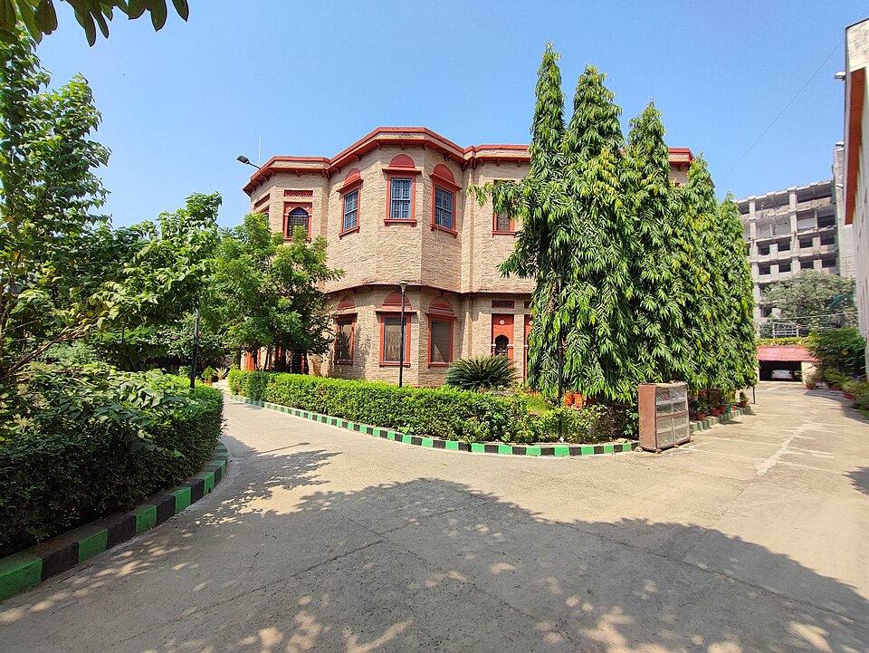 पटना का ख़ुदाबख़्श ओरिएंटल पब्लिक लाइब्रेरी — दो मंज़िला ईंट की इमारत, जिसकी मेहराबदार खिड़कियों के घेरे लाल रंग के हैं और एक कोणीय झरोखा है; वह छँटी हुई झाड़ियों और आम जैसे पेड़ों के पीछे, पक्के रास्ते के छोर पर, साफ़ नीले आकाश तले खड़ी है
