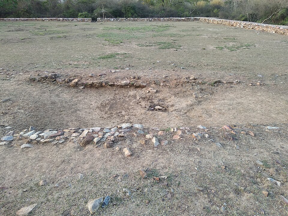 The site called Bimbisara's Jail at Rajgir — low lines of rough undressed stone footings marking out rectangular rooms on bare dusty ground, with a rebuilt rubble boundary wall and scrub-covered hill slope along the far edge