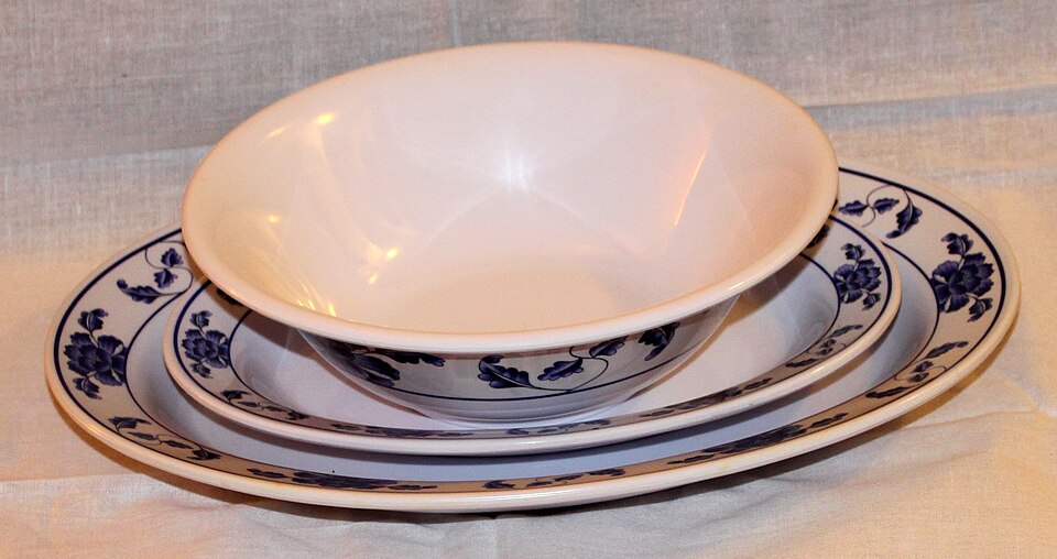 A nested set of glossy white melamine tableware on a cream cloth — two plates of different sizes and a deep bowl, each with a blue floral border, showing the hard smooth sheen of moulded melamine crockery