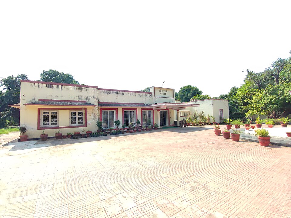 A long single-storey cream building with red-painted window frames and a small Hindi signboard reading Rajendra Smriti Sangrahalaya, potted plants lined along its verandah and a wide brick-paved forecourt in front, trees closing in behind — the memorial museum inside the Sadaqat Ashram campus in Patna