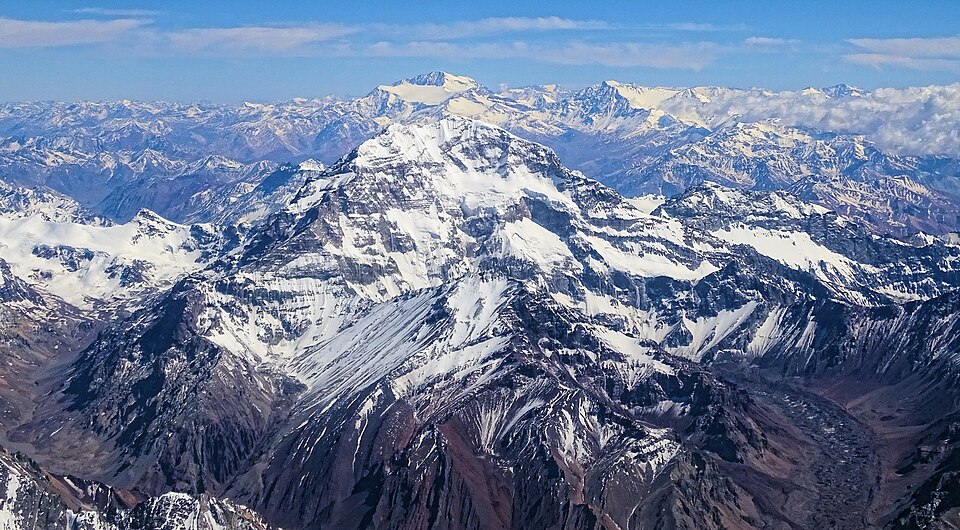 Aconcagua from the air — a broad snow-plastered massif of dark rock ribs and hanging glaciers standing well above every surrounding ridge, with rank after rank of brown Andean crests running to the horizon on either side