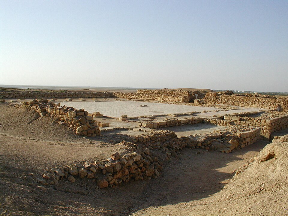 The excavated ruins at Banbhore in Sindh — low courses of rough sandstone walls marking out rooms and a colonnade around a flat paved courtyard, standing on bare sun-baked ground with the flat coastal creeklands stretching to the horizon behind