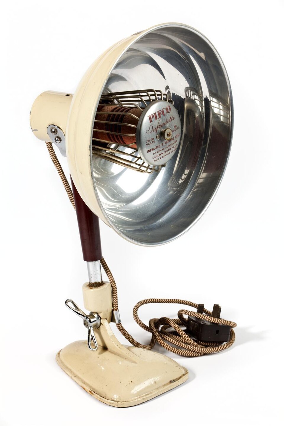 An infra-red therapy lamp photographed against a white background — a large polished chrome reflector dish on a tilting cream stand, a coiled heating element behind a wire guard at its centre carrying a maker's plate reading 'INFRA-RED & RADIANT HEAT', with a braided flex and plug coiled beside the base