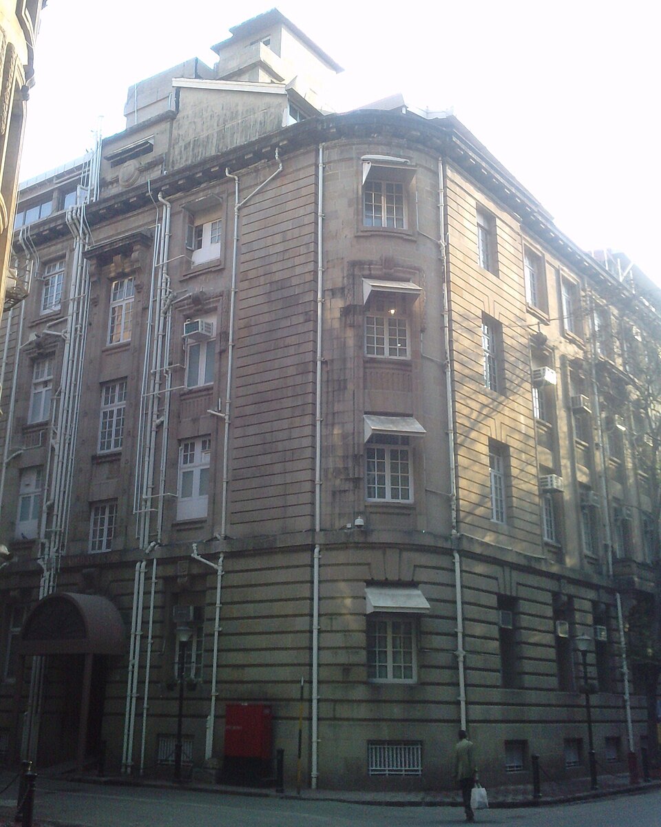 Bombay House seen from the street corner in Fort, Mumbai — a four-storey building of coursed grey-brown stone with a rounded corner bay, projecting oriel windows with awnings, external service pipes running up the facade and a canopied entrance at street level
