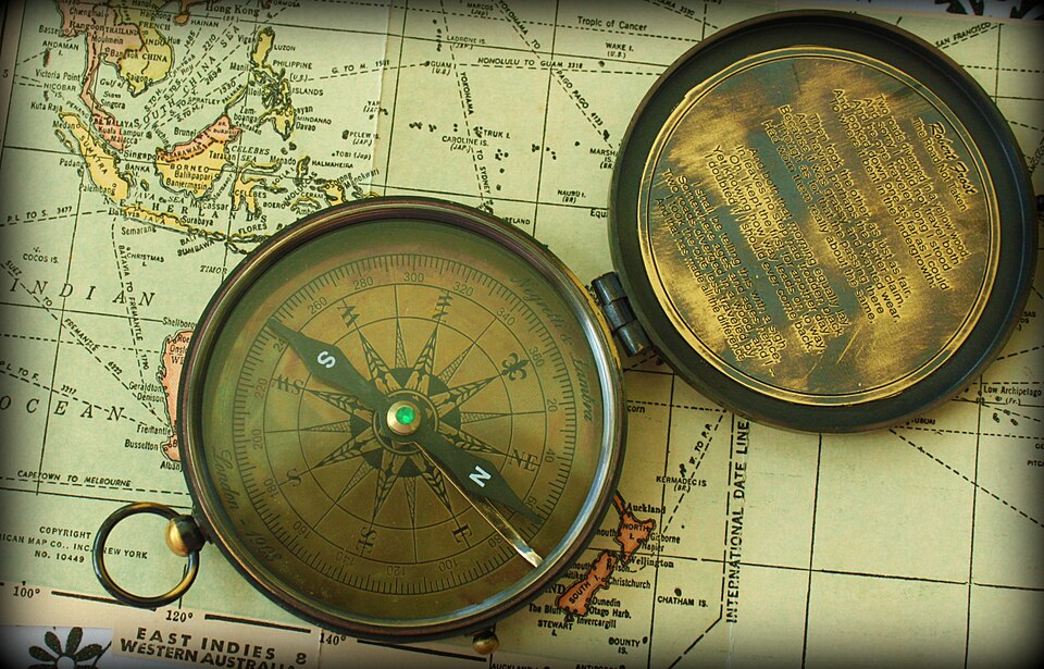 An open brass magnetic pocket compass resting on an old world map, its printed card showing a compass rose with cardinal and intercardinal letters and a degree ring, with the blue-tipped needle settled over the card