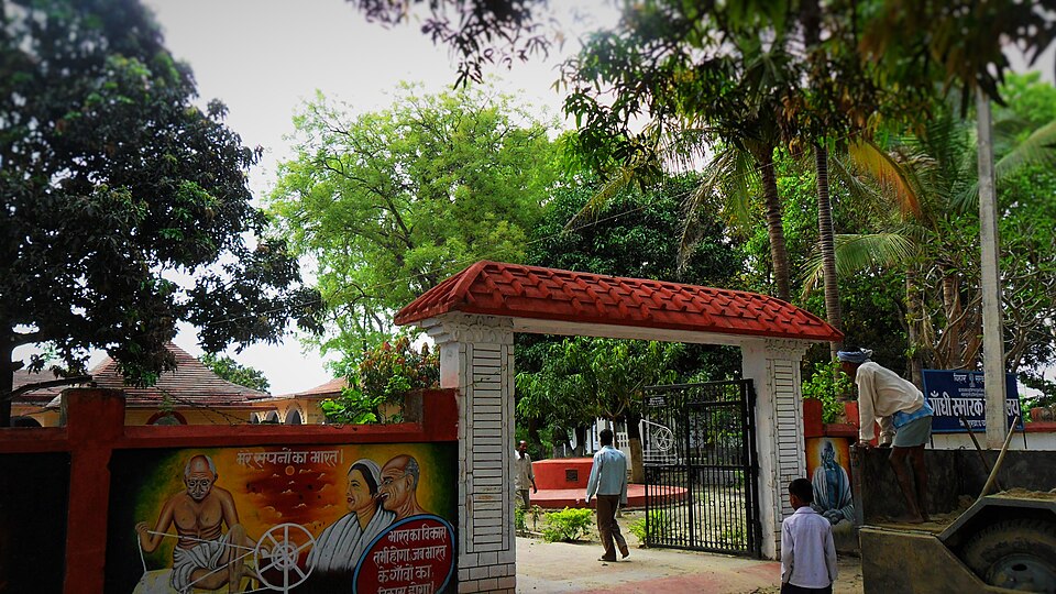 The tiled gateway of Bhitiharwa Gandhi Ashram in West Champaran, its boundary wall painted with a mural of Gandhi at a spinning wheel beside a Hindi signboard, with the ashram buildings and trees behind