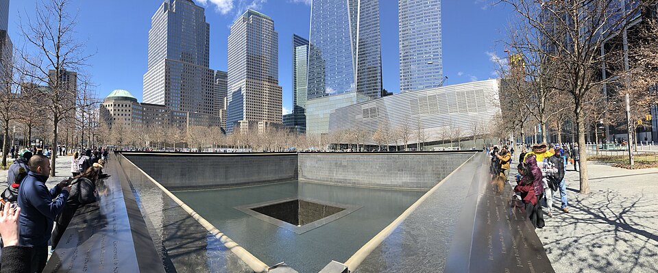 One of the two square reflecting pools of the National September 11 Memorial in Lower Manhattan, water falling down its granite walls into a central void, ringed by a bronze parapet of engraved names with visitors around it and the World Trade Center towers behind