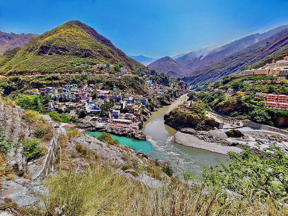 The confluence at Devprayag seen from above, the turquoise Bhagirathi meeting the muddy brown Alaknanda at a sharp angle below the terraced hillside town, the combined stream flowing away as the Ganga