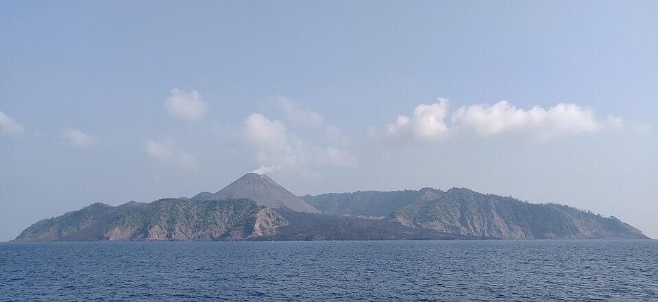 Barren Island seen from the sea, a dark grey cinder cone trailing a wisp of steam above a breached caldera rim, with a bare lava field running down to the shoreline between green flanks