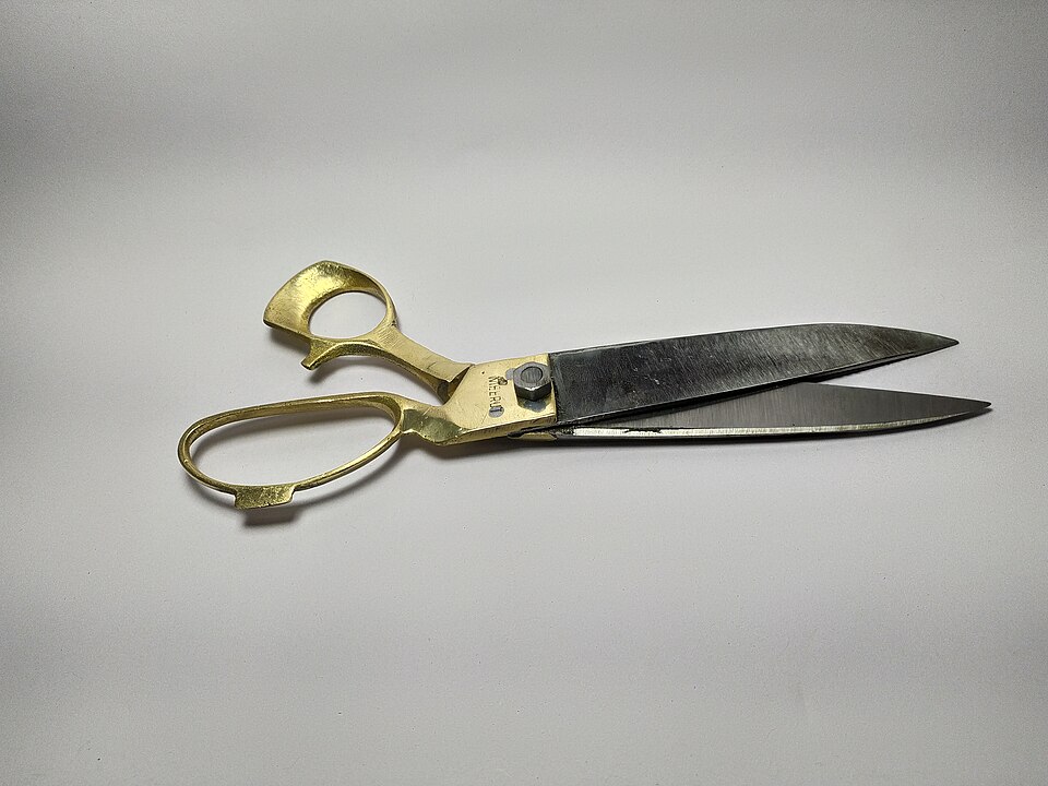 A pair of hand-forged tailor's scissors from Meerut lying open on a plain grey surface, brass bow handles riveted to long polished steel blades