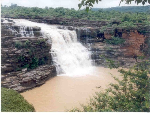 Karkat waterfall on the Karmanasa, a broad white sheet of water dropping over the layered sandstone lip of the Kaimur escarpment into a muddy plunge pool, with scrub forest on the plateau above