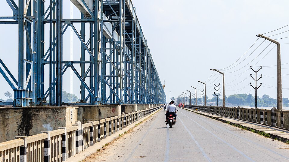 The road deck of the Kosi (Koshi) Barrage running between its long line of blue steel gate-hoist gantries, with a motorcyclist crossing and the river visible on the right