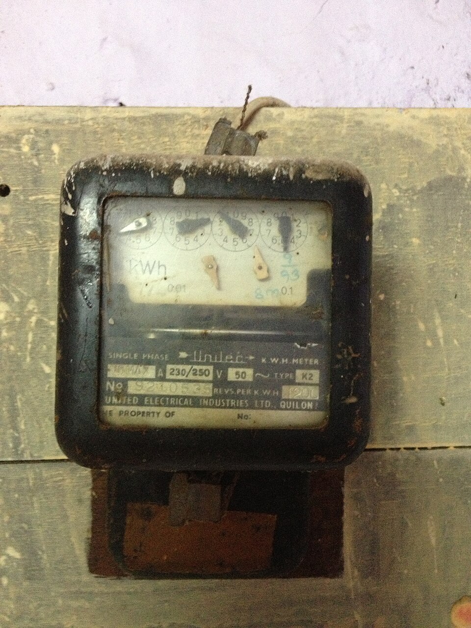 A single-phase domestic electricity meter screwed to a wooden board on a wall, its four-dial register marked kWh above a plate reading SINGLE PHASE K.W.H. METER, 230/250 V, 50 Hz