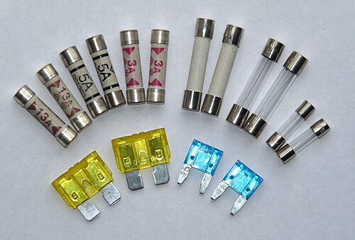 An assortment of electrical fuses laid out on a white surface: ceramic cartridge fuses marked 13A, 5A and 3A with metal end-caps, clear glass cartridge fuses with the thin fuse element visible inside, and coloured blade fuses