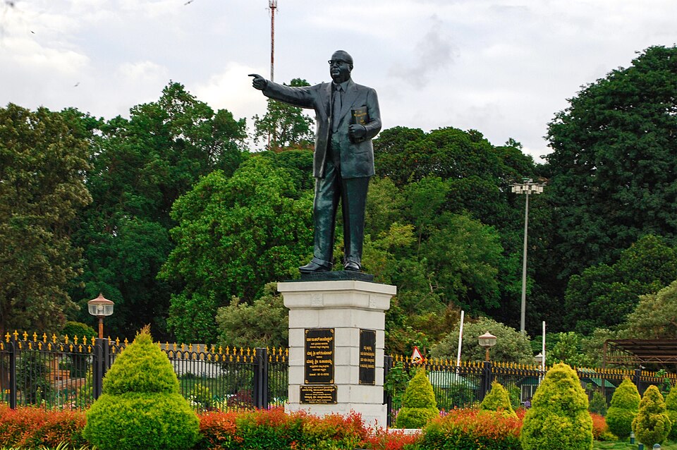 A bronze public statue of Dr B. R. Ambedkar in a suit on a stone pedestal, holding a bound copy of the Constitution in one hand and pointing ahead with the other, set in a garden