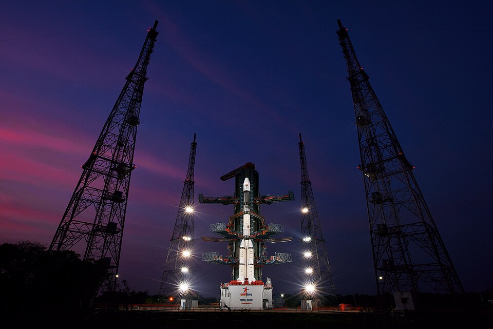 A GSLV launch vehicle standing floodlit on the Second Launch Pad at Satish Dhawan Space Centre SHAR, Sriharikota, at dusk, flanked by two tall lattice lightning-protection towers