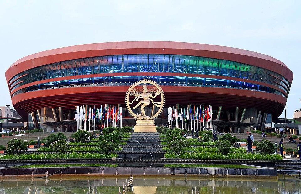 The elliptical copper-red facade of the Bharat Mandapam convention centre at Pragati Maidan, New Delhi, with the golden Nataraja statue and rows of national flags on the forecourt