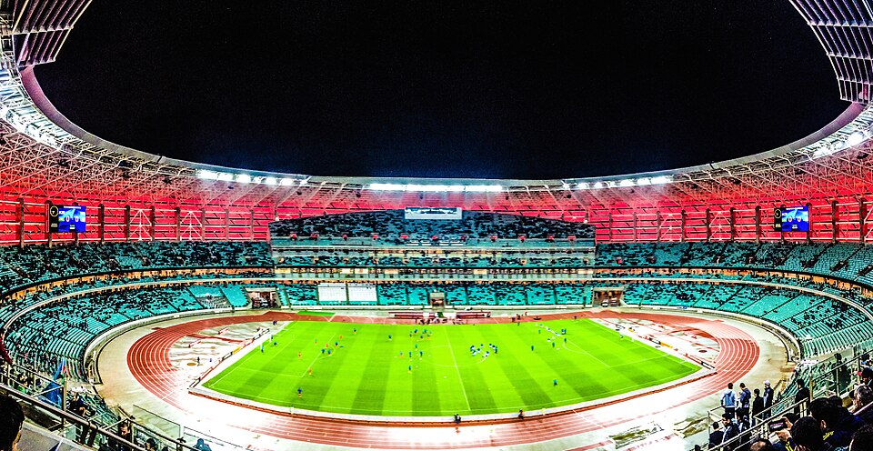 Night interior panorama of the Baku Olympic Stadium showing its red and turquoise tiered seating bowl, floodlit running track and green pitch