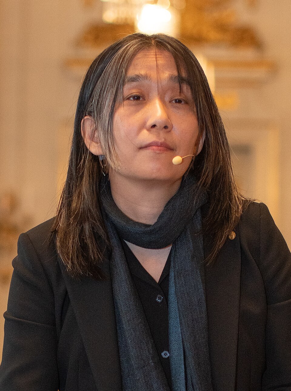 Portrait of the South Korean novelist Han Kang in a dark jacket and scarf, wearing a headset microphone, photographed during Nobel week 2024