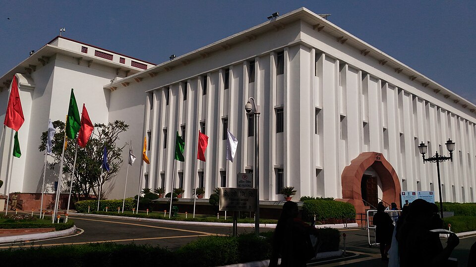 Exterior of Vigyan Bhawan on Maulana Azad Road, New Delhi: a long white building faced with vertical fluted piers, with a red-sandstone arched entrance porch and a row of flagpoles in front