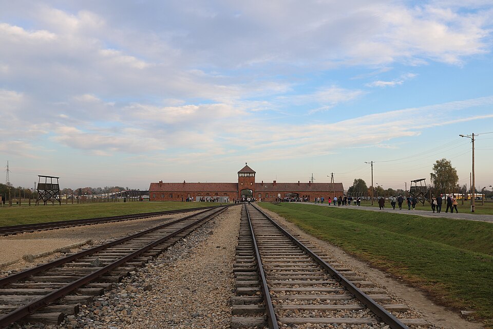The red-brick main gatehouse of Auschwitz II-Birkenau seen straight down the railway tracks that run through its central archway, with watchtowers on either side of the camp field