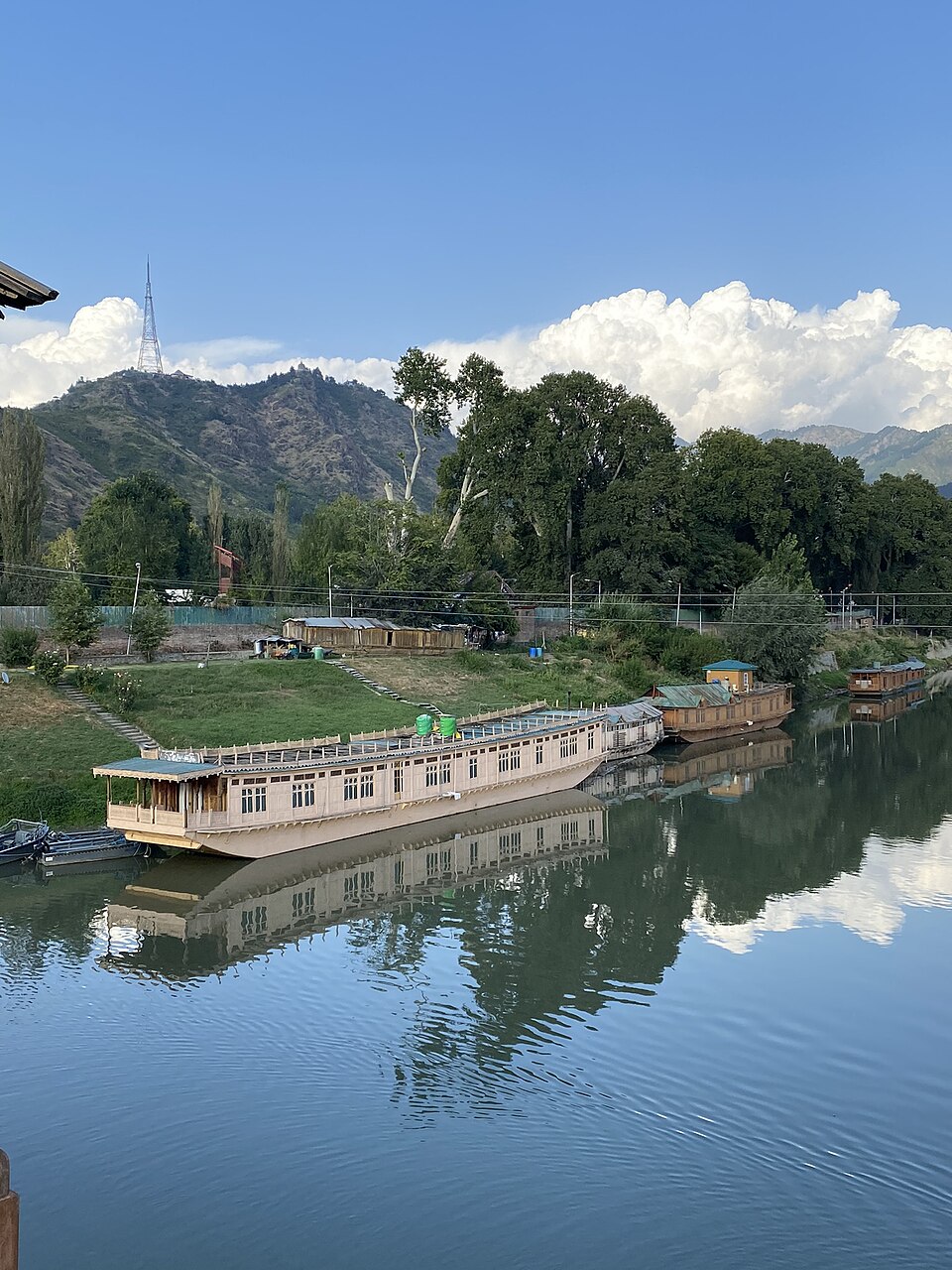 The Jhelum river at Srinagar seen from Zero Bridge, with wooden houseboats moored along the bank and the Shankaracharya hill and its transmission tower rising behind